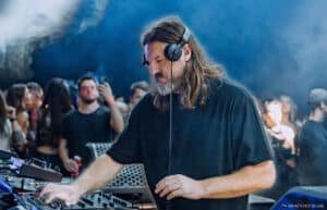 A DJ with long hair and a beard, wearing headphones, is intensely focused on mixing music at an electronic music event with a crowd in the background.