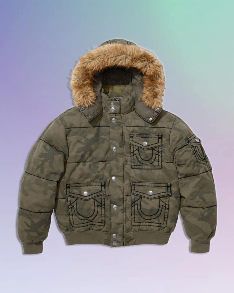 A camo-patterned puffer jacket with a fur-lined hood and multiple buttoned pockets, set against a gradient background. Part of the Supreme x True Religion collab.