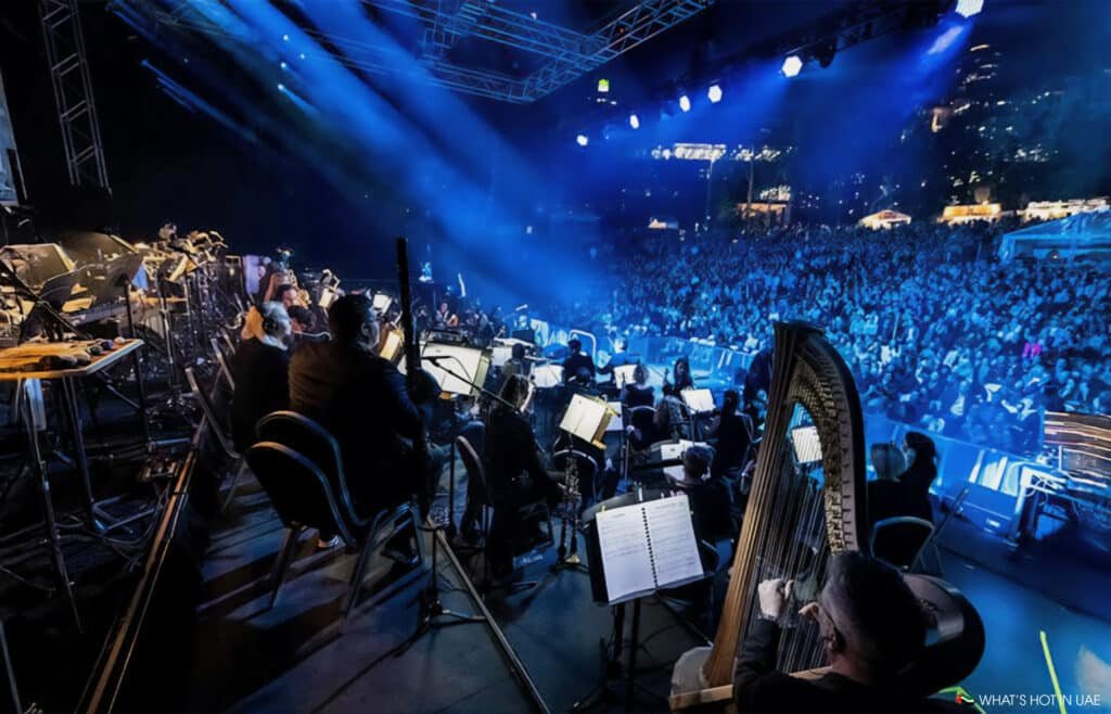 View from behind the Synthony orchestra performing on stage with a large, illuminated audience in the background.