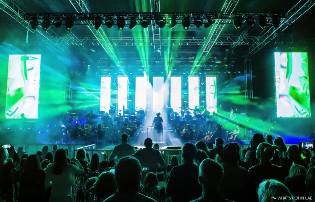 A vibrant concert scene with a conductor and orchestra on stage, illuminated by green and blue lights, while a crowd watches in silhouette.