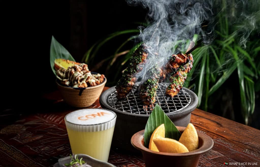 Grilled skewers with herbs on a round charcoal grill, accompanied by a dish of golden wedges and a creamy cocktail with 'COYA' written on its foam, set on a wooden table with green plants in the background.