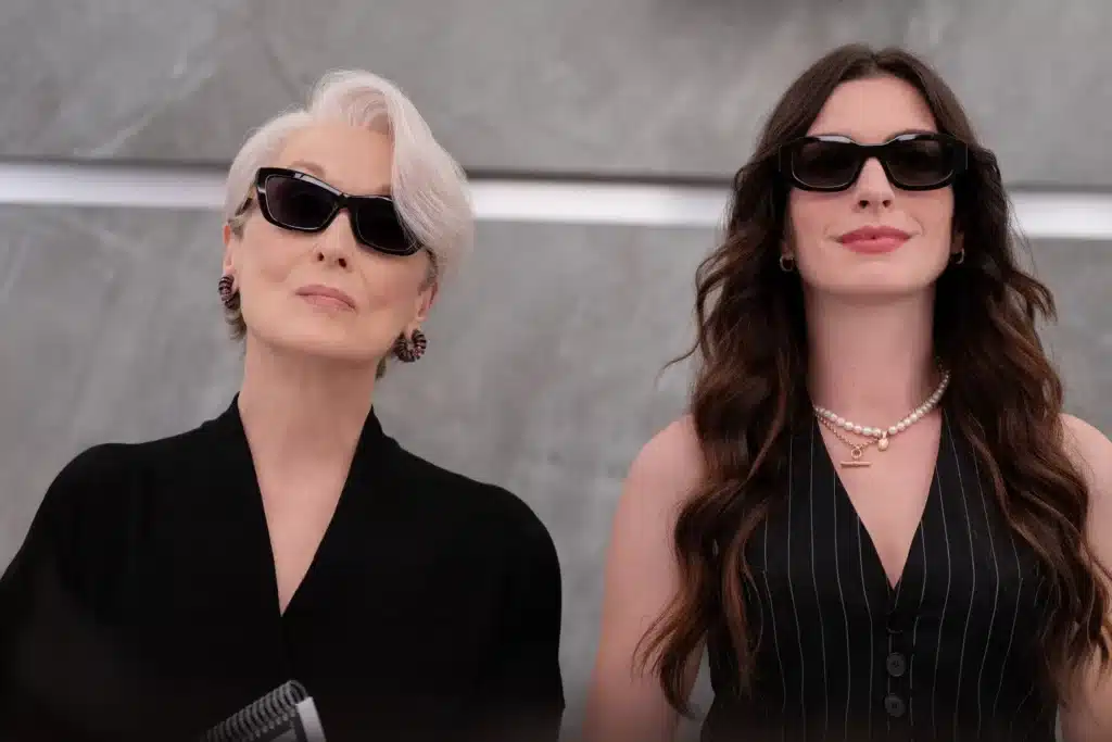 Two women wearing sunglasses and stylish outfits stand side by side against a marble background. One has short white hair and the other has long wavy brown hair.