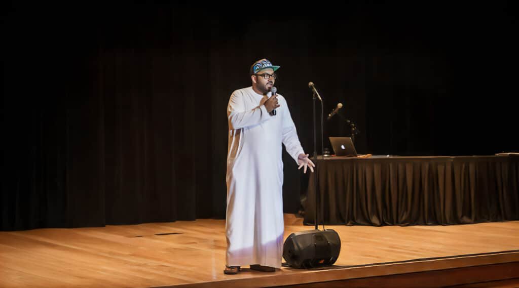 Ali Al Sayed wearing a traditional white thobe and a cap is speaking into a microphone on stage. There is a table with a laptop and two microphones in the background. The stage has wooden flooring and a black curtain backdrop.