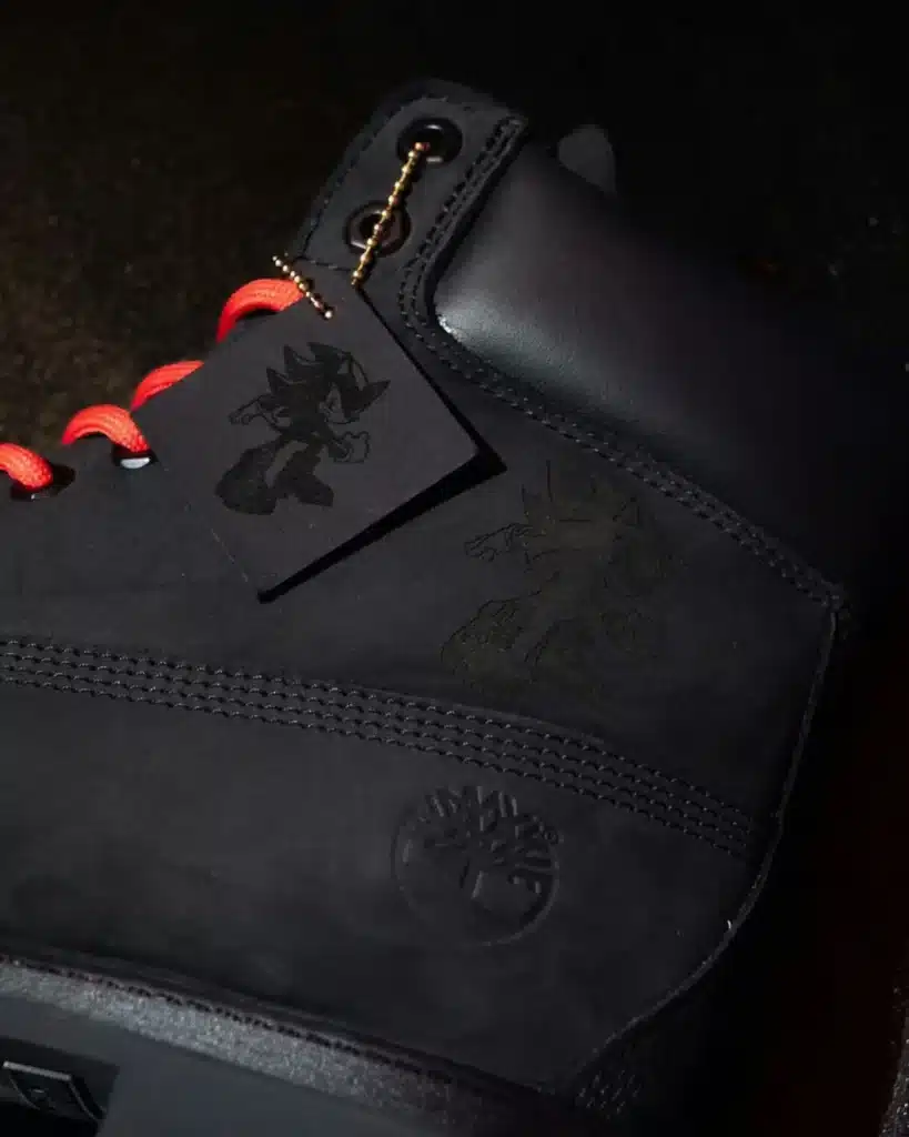 A close-up of black Timberland Shadow the Hedgehog Boots featuring red laces and a tag with a character illustration. The boot has a Timberland logo and an engraved character image on the side.