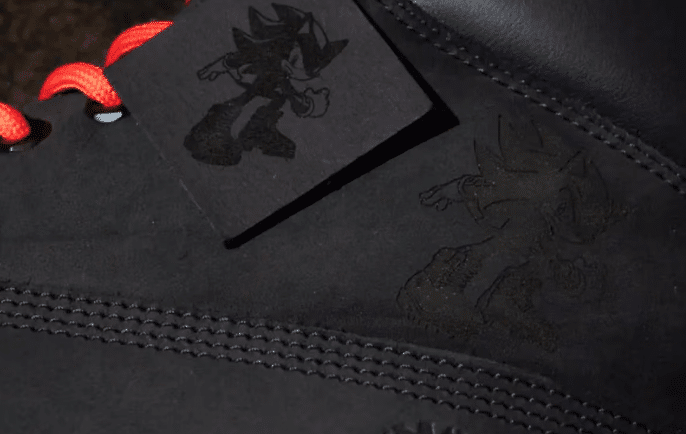 Close-up of a black sneaker with red laces displaying an embossed image of a cartoon hedgehog and a matching tag, both in a shadowy dark tone.