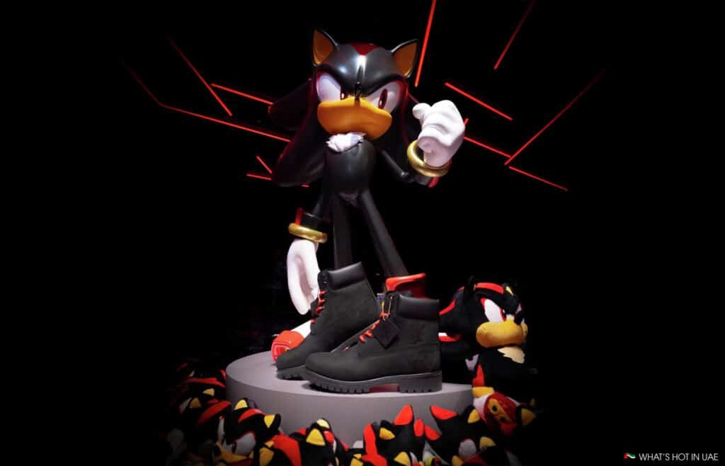 A display featuring a statue of a black and red cartoon hedgehog character standing on a pedestal, surrounded by plush toys of the same character. In front of the statue are a pair of black boots with red accents.
