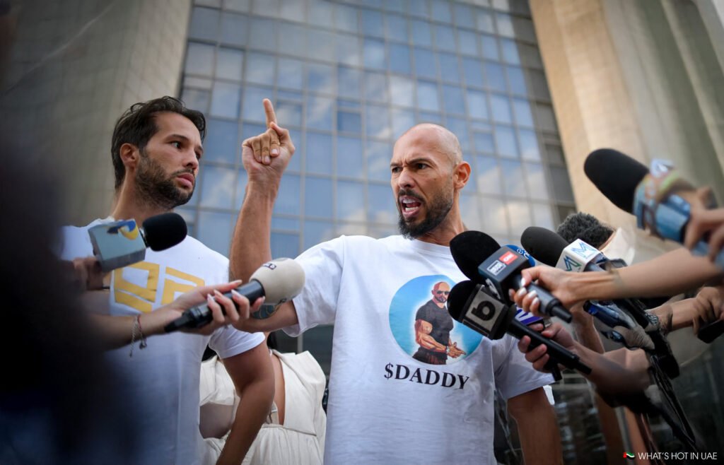 Andrew Tate vs Chase DeMoor: Tristan and Andrew Tate are surrounded by microphones from various media outlets in front of a modern building, with one man gesturing emphatically while wearing a shirt with a graphic of a muscular figure labeled '$DADDY.'