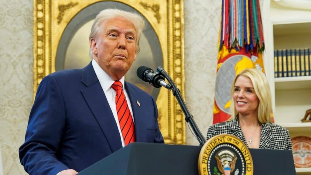 A man in a suit and red tie stands at a podium with a seal that reads "The President of the United States," while a woman in a checkered blazer stands beside him smiling. The setting is an ornate room with portraits and flags in the background.