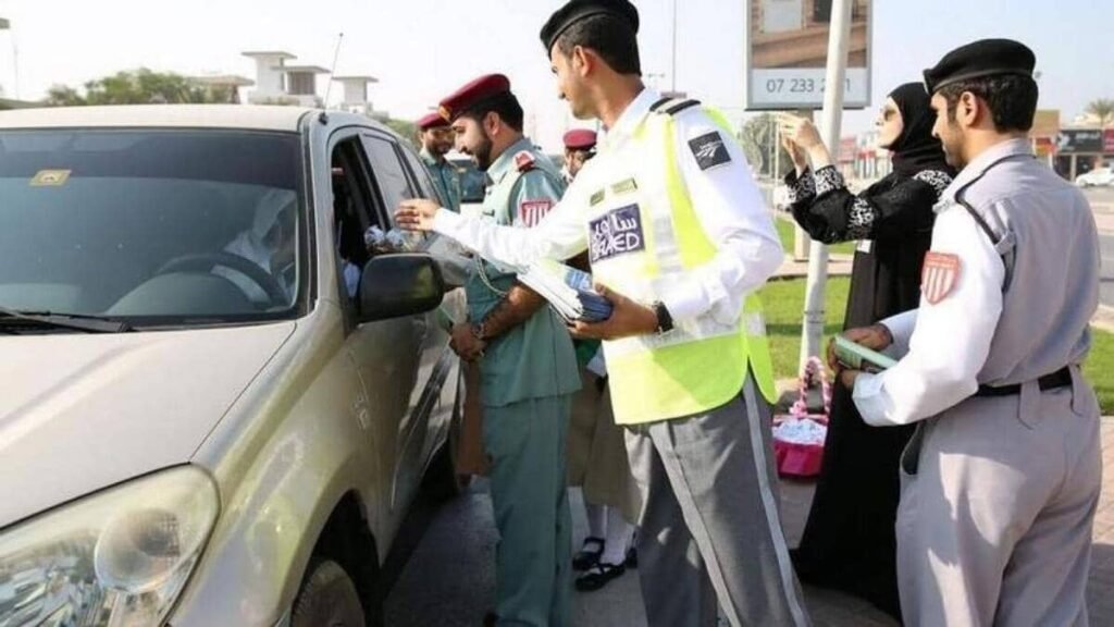 A group of people in uniform, possibly police officers, distribute flyers to motorists through car windows on a street. A woman wearing a hijab is assisting them. A beige car is in the foreground.