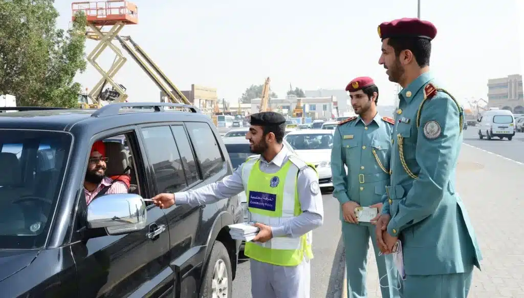 A community police officer hands a UAE Fine to a driver in a black SUV, while two uniformed police officers stand nearby on a busy street.