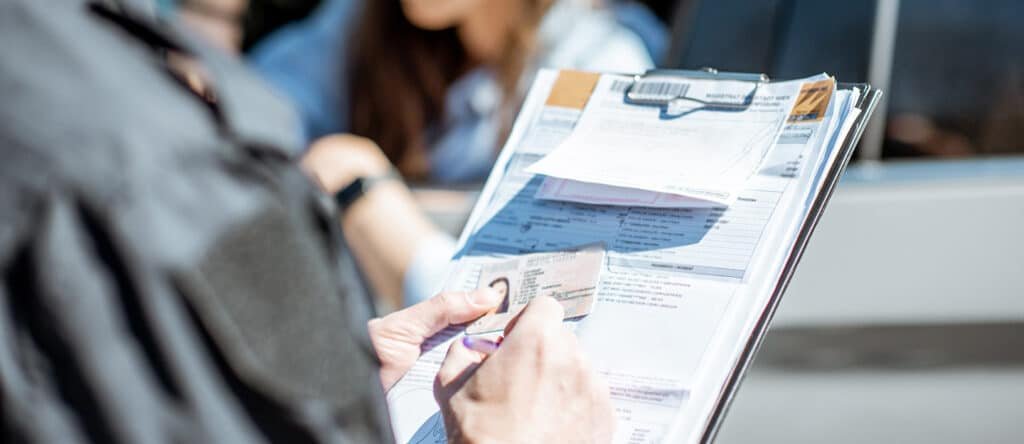 A person holding a driver's license and writing on a clipboard filled with various UAE Fine documents, with a blurred background of another person near a car window.