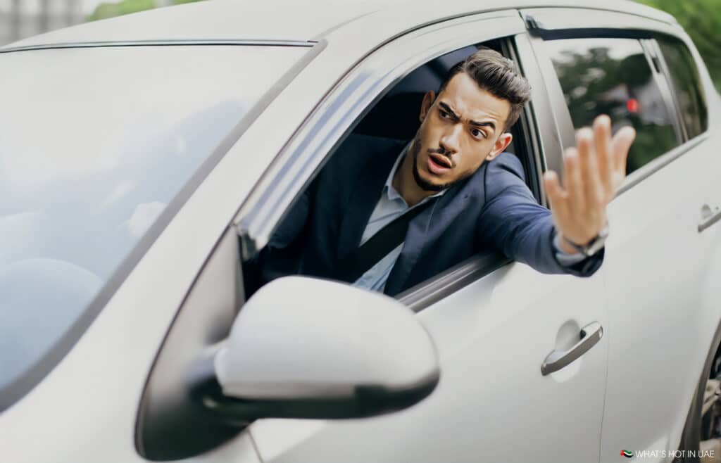 A man in a suit is sitting in a white car with the window rolled down, gesturing with his hand and appearing frustrated or confused.
