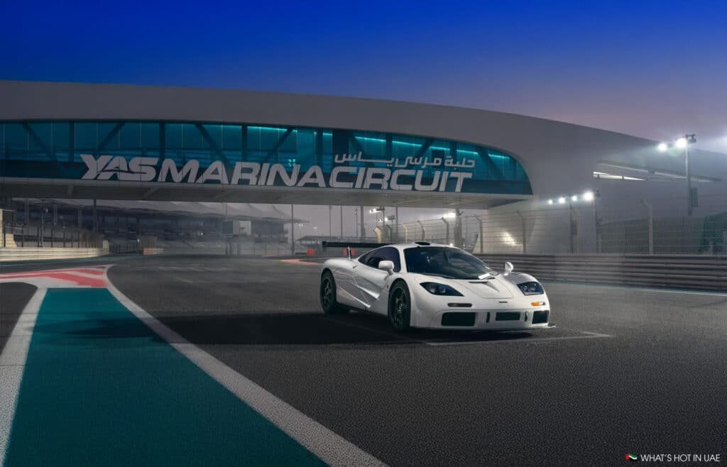 A white sports car parked on the track at Yas Marina Circuit, with the circuit's bridge and signage visible in the background, illuminated by floodlights at dusk.
