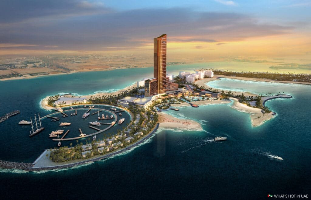 Aerial view of a coastal resort featuring a tall, modern skyscraper surrounded by luxurious buildings, palm trees, and a marina with several yachts docked. The resort is set against a backdrop of turquoise waters and sandy beaches with a cityscape visible in the distance under a sunset sky.