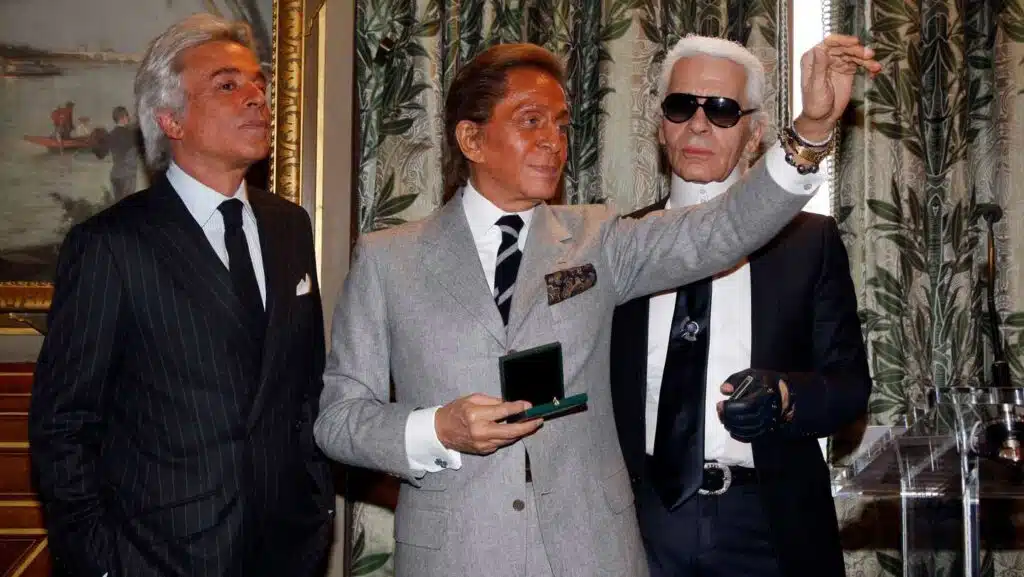 The History of Valentino: Three men in formal suits stand together, with the man in the centre holding a small green box. They are in a decorated room with a painting and patterned curtains behind them.