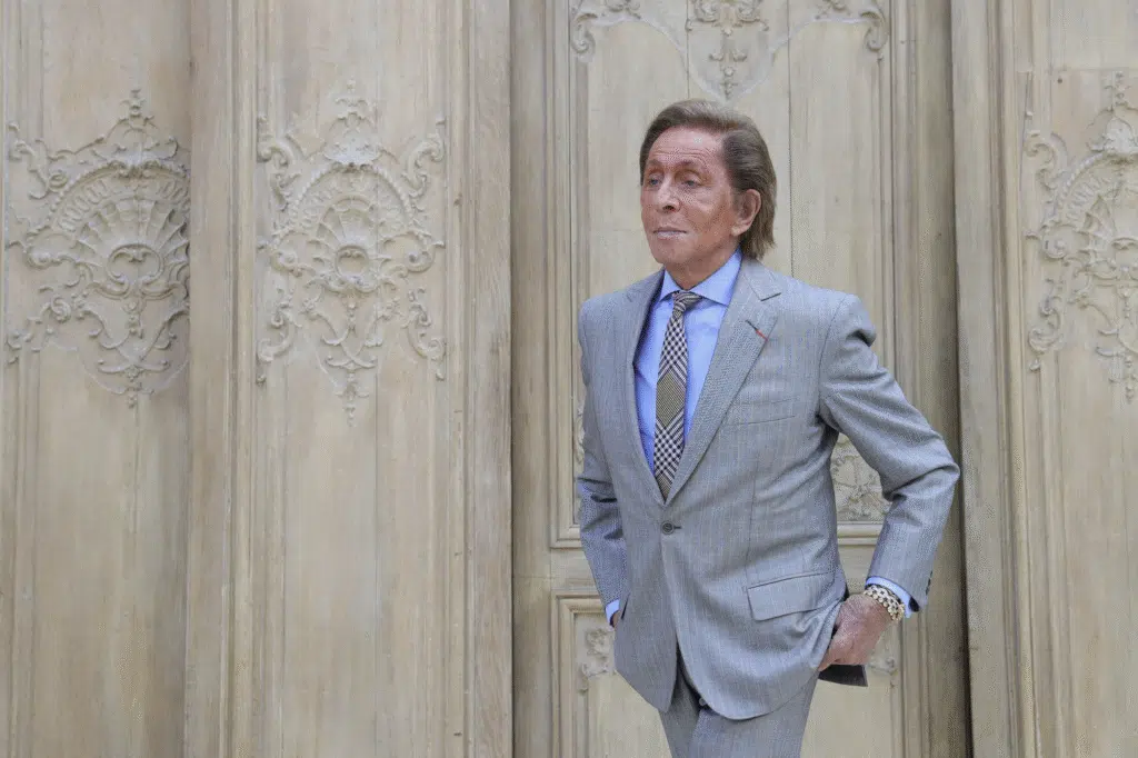 A man in a light gray suit with a blue shirt and patterned tie stands against an ornate, intricately carved wooden wall.