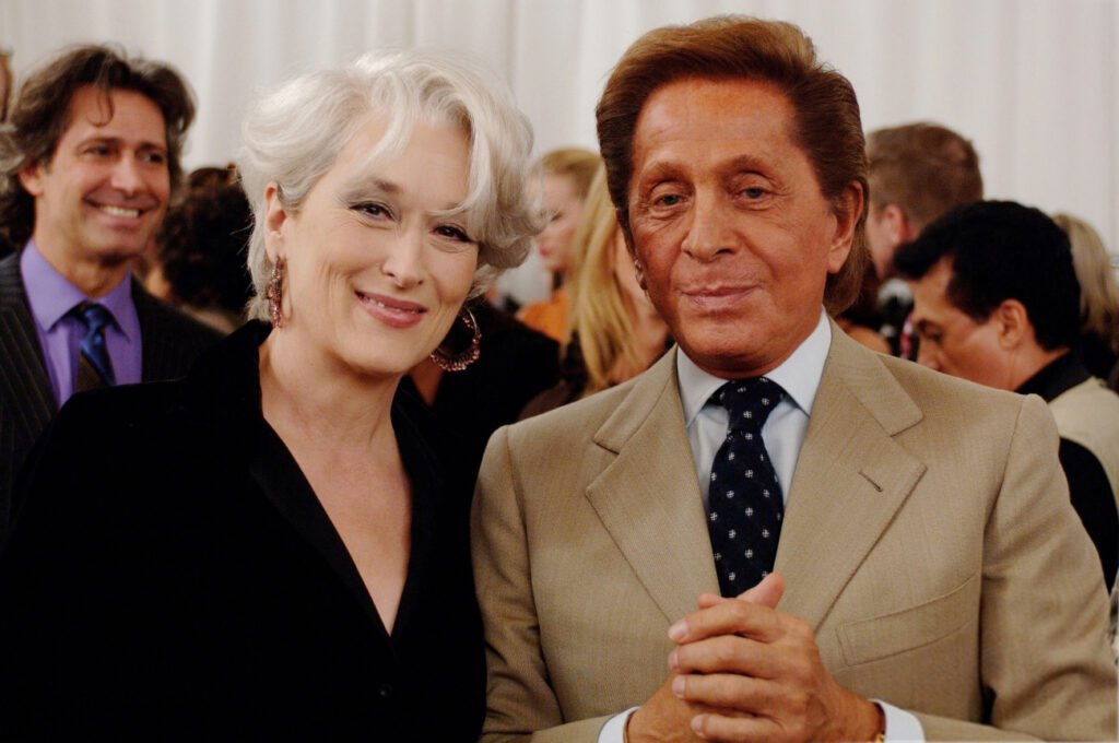 Meryl Streep with short, white hair in a black outfit stands next to Valentino with brown hair in a light tan suit and a polka dot tie, both smiling at a social event.