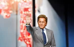 A man in a gray suit and blue polka dot tie waves with his right hand. The background features a blurry pink and red floral arrangement.