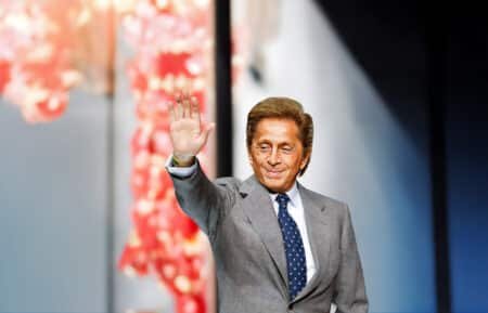 A man in a gray suit and blue polka dot tie waves with his right hand. The background features a blurry pink and red floral arrangement.