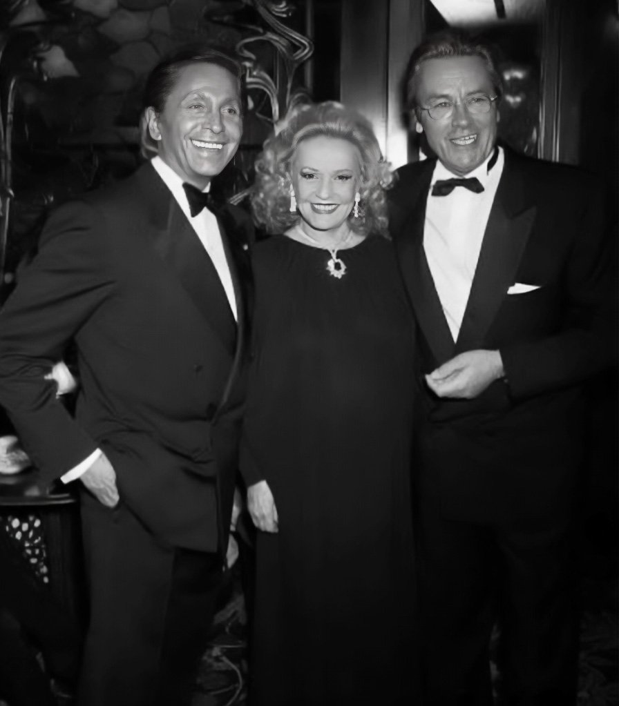 The History of Valentino: Three people dressed in formal evening wear, standing closely together and smiling for a photo, with two men in tuxedos and a woman in a dark dress adorned with jewelry.