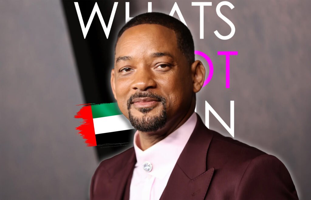 A person wearing a burgundy suit and a pink shirt with a crystal button, standing in front of a blurred background with the word 'WHAT'S' visible behind them. A stylized United Arab Emirates flag is placed on the image.