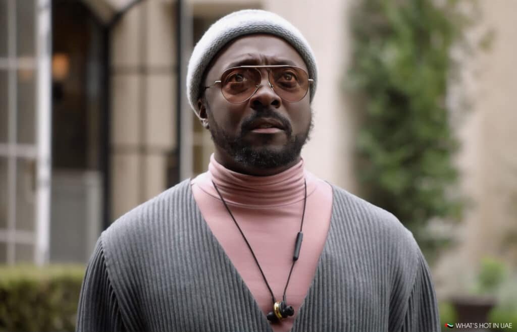 Will.i.am wearing a gray beanie, round sunglasses, a pink high-neck sweater, and a ribbed gray cardigan. He is outdoors with blurred greenery in the background.