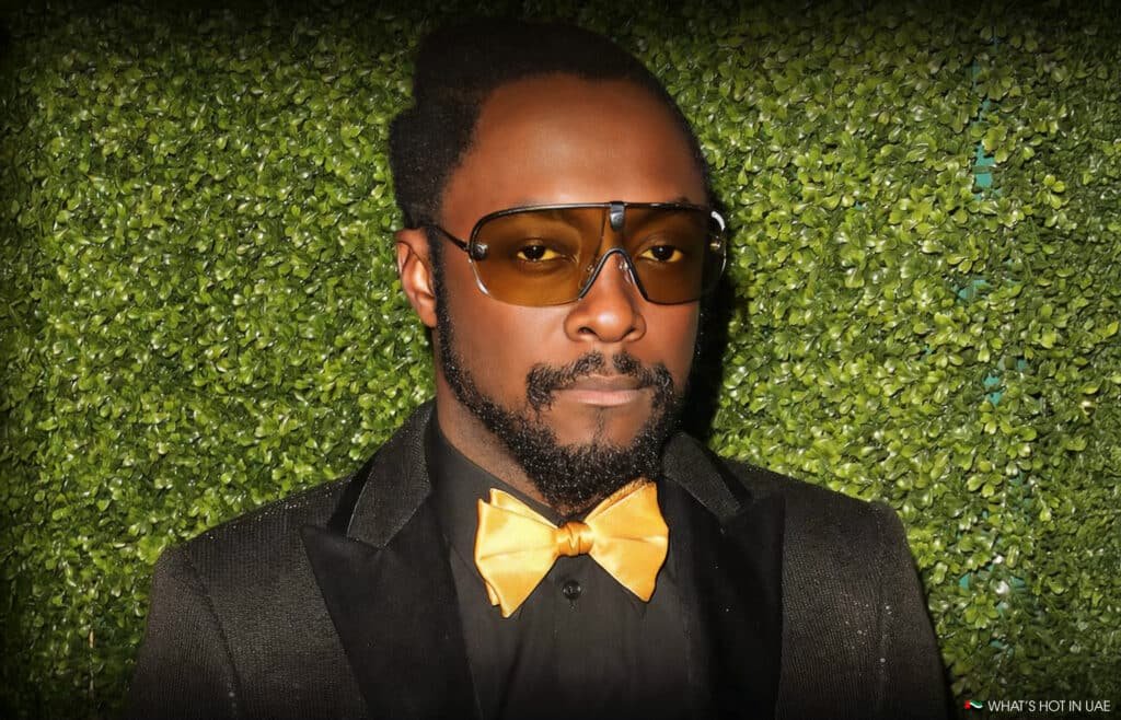 Will.i.am with a beard and mustache wearing yellow-tinted sunglasses, a yellow bow tie, and a black suit stands in front of a wall of greenery.