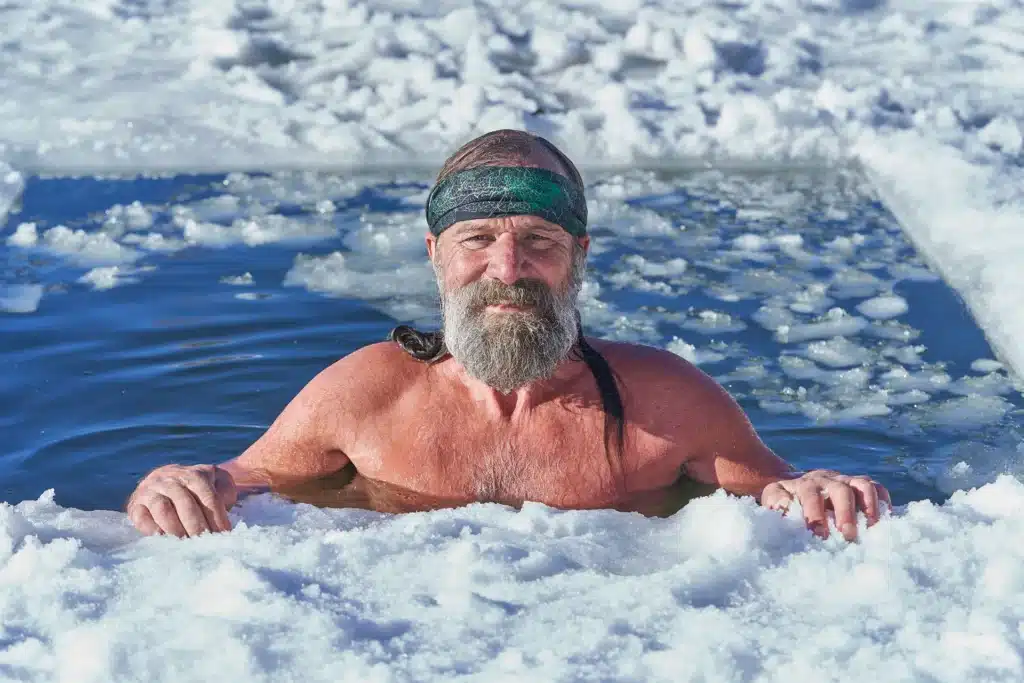 Wim Hof with a beard and headband immerses himself in a pool of icy water, surrounded by snow.
