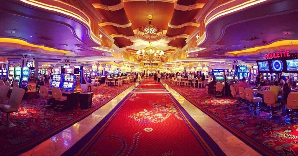 Interior of a casino with rows of slot machines and a central red carpet walkway, under a decorative ceiling with chandeliers.