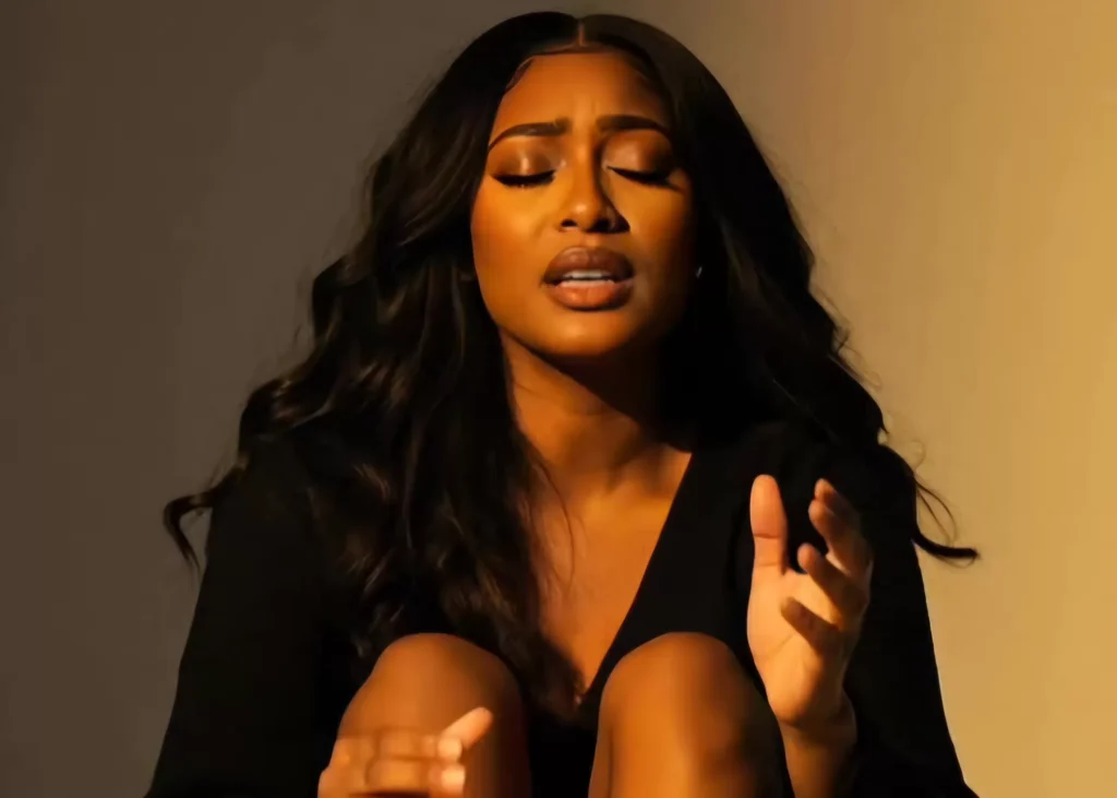 A woman with long wavy hair is sitting with her knees up, eyes closed, and hands raised, appearing to be in a moment of emotion.