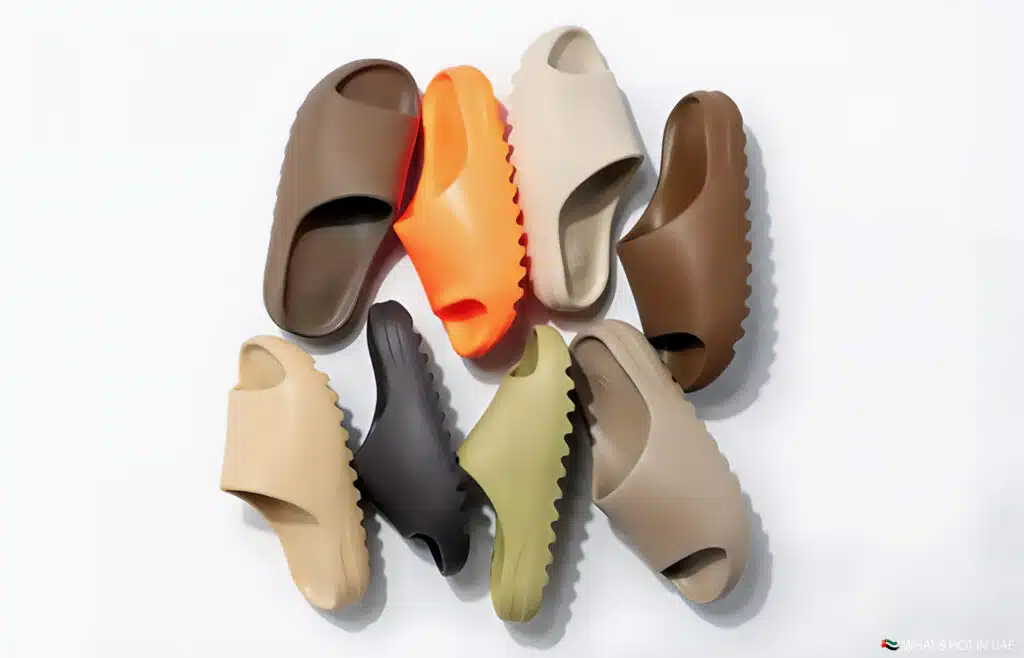 A collection of Kanye West Yeezy Slides in various colors, including brown, beige, orange, and green, arranged in a circular pattern on a white background.