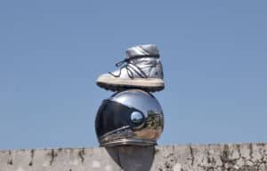 A silver boot sits atop a reflective, chrome astronaut helmet on a concrete ledge against a clear blue sky.