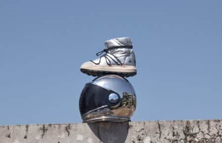 A silver boot sits atop a reflective, chrome astronaut helmet on a concrete ledge against a clear blue sky.