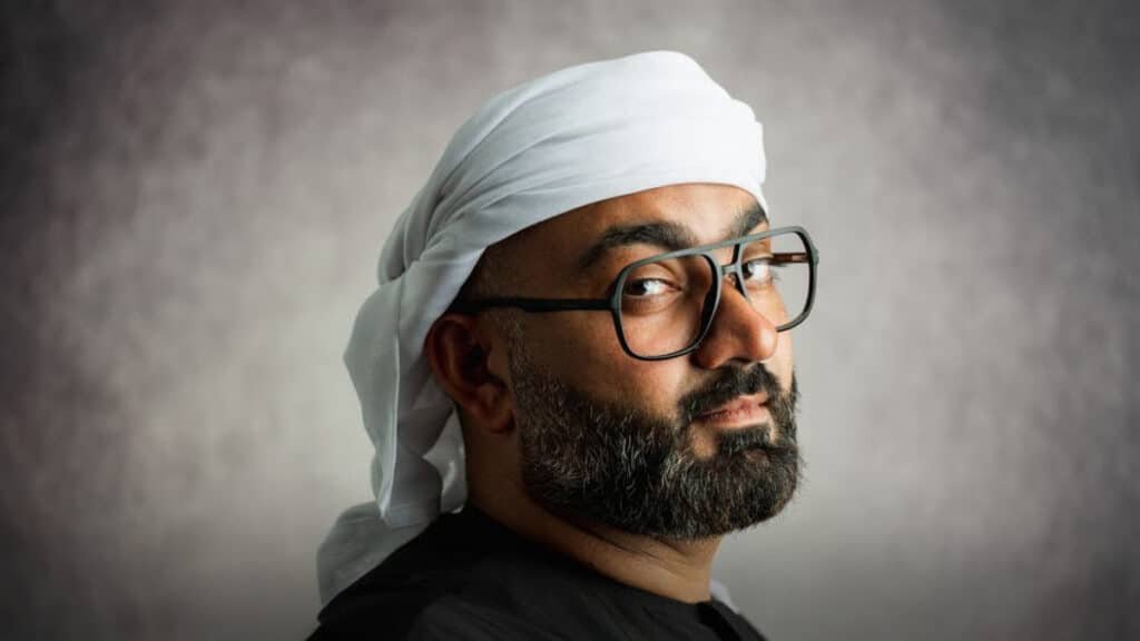 A bearded man wearing glasses and a traditional white headscarf, tilting his head slightly back, against a neutral background.