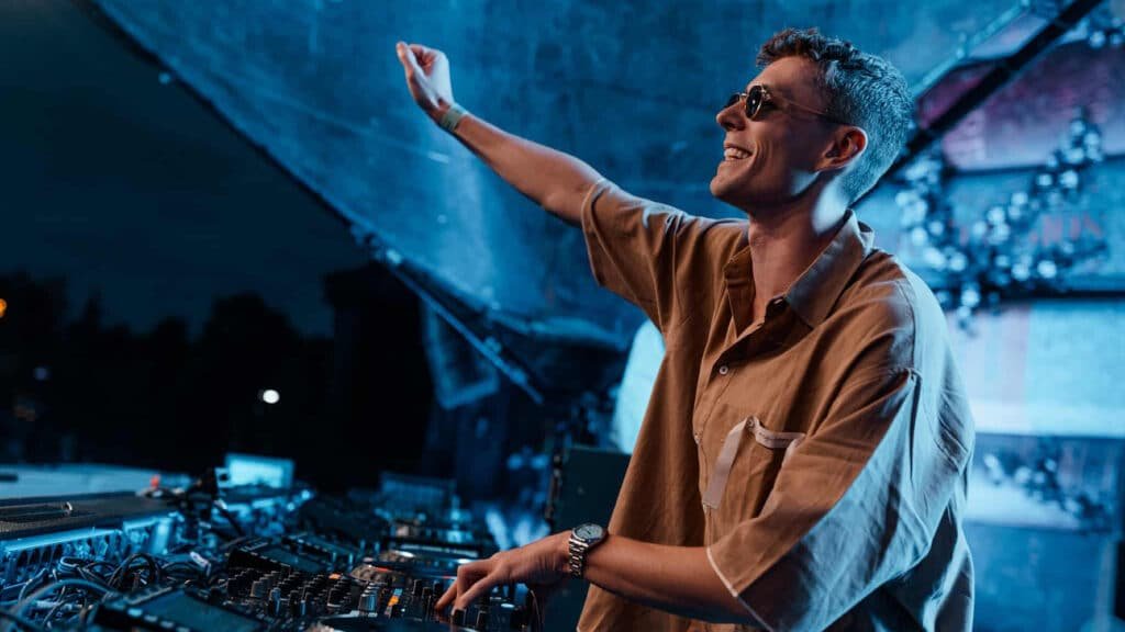 Lost Frequencies wearing sunglasses and a brown shirt is performing at a music event, with one hand on the mixing console and the other raised in the air. The scene is illuminated with blue lighting.