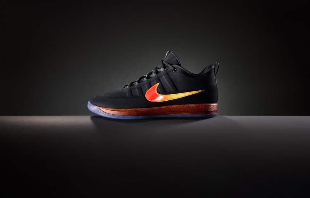 A black sneaker with a red and orange gradient Nike swoosh and a translucent sole on a dark background.