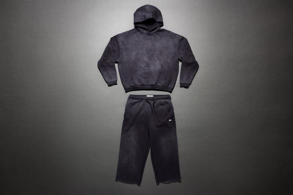 A dark gray hoodie and sweatpants set laid out on a gray background, featuring a minimalist design with subtle branding.