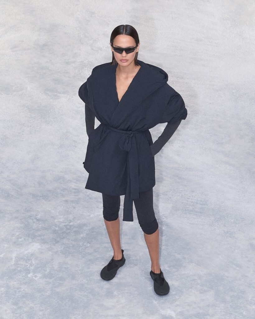 NikeSKIMS Drop 2 Collection: A person wearing a stylish, dark-colored outfit consisting of a belted coat, leggings, and slip-on shoes, standing on a light gray textured surface. They have sleek hair, and are accessorized with narrow sunglasses, exuding a modern and chic look.