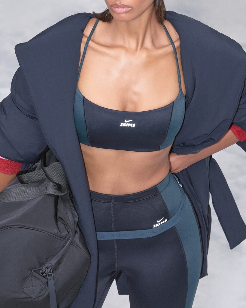 NikeSKIMS Drop 2 Collection: A person wearing a navy blue sports bra and leggings with "SKIMS" branding, layered with a loose dark jacket and carrying a gym bag.