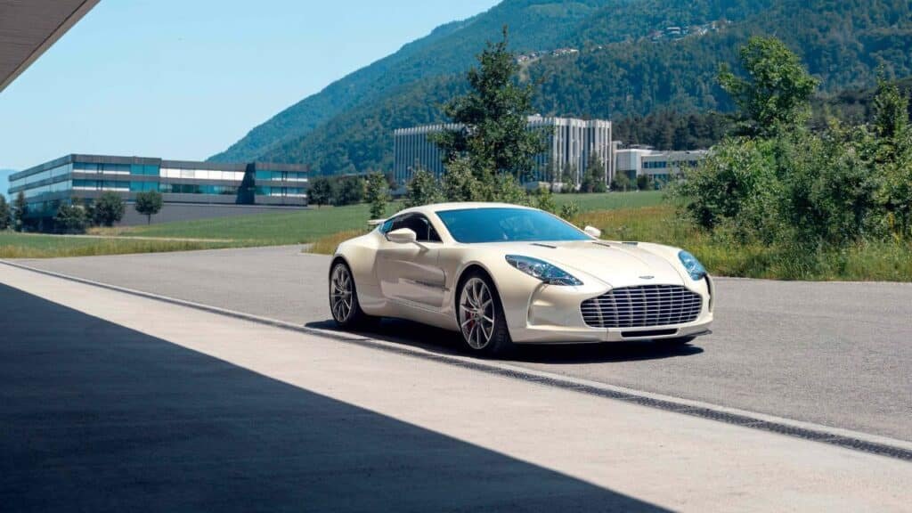 A sleek white Sotheby’s sports car parked on a road with modern buildings and lush green hills in the background.