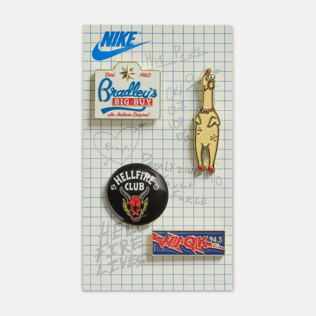 A set of four enamel pins on a grid-patterned card featuring a Nike logo. The pins include a retro-style store sign reading "Bradley's Big Buy," a cartoon chicken, a black pin with "Hellfire Club" and a devil face, and a logo pin for "WSJQK 94.5 FM."