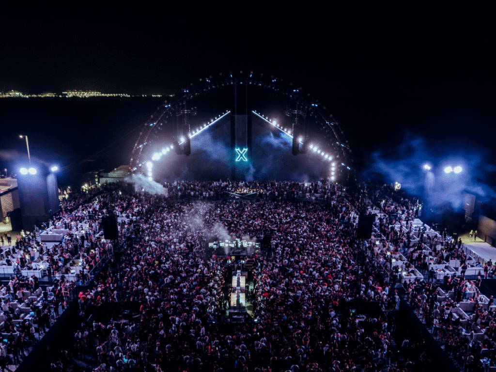 A large outdoor music festival at night with a crowd of people in front of a brightly lit stage featuring a large "X" symbol. The scene is surrounded by smoke and colorful lights, creating a vibrant atmosphere.