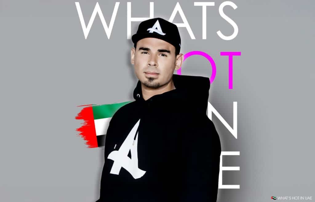 A person wearing a black hoodie and cap with a white 'A' logo, standing against a gray background with partial text in large letters. The image also features a digitally painted flag of the United Arab Emirates.