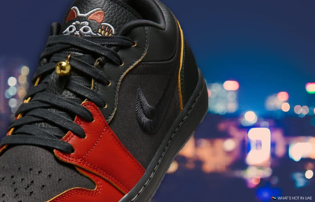 A black Air Jordan 1 Low 'Money Cat' sneaker with red accents and a rat emblem on the tongue, set against a blurred cityscape backdrop at night.
