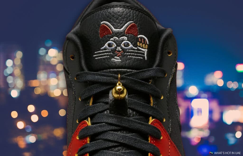 A sneaker with a cat design on the tongue, featuring red highlights and a small bell, set against a blurred cityscape.