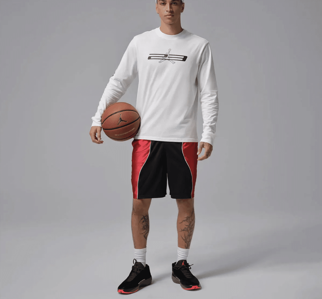 A person holding a basketball, wearing a white long-sleeve shirt with a graphic design, black and red shorts, and black Air Jordan 40 'Infrared' sneakers with red details.