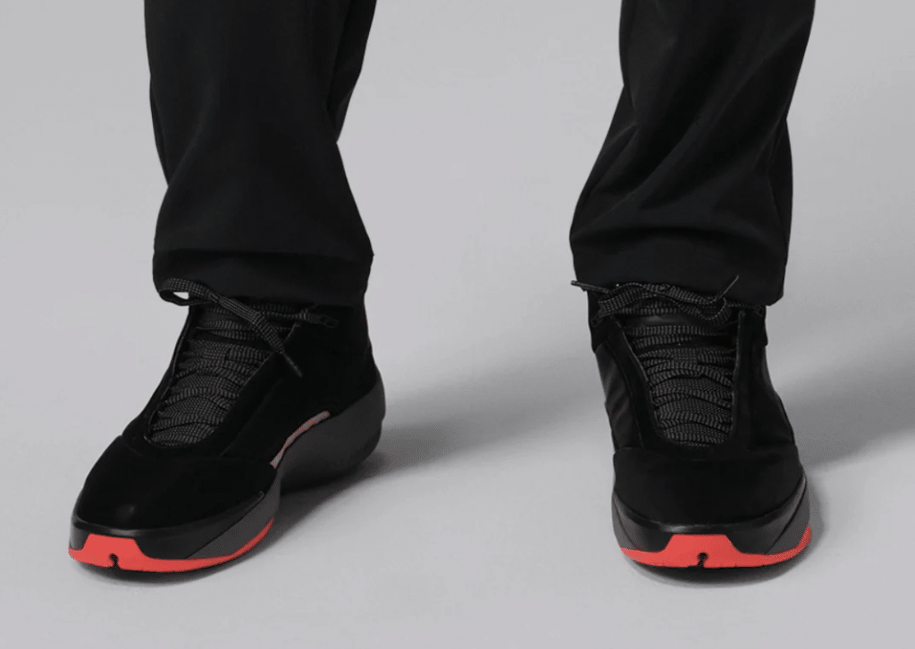 A pair of black sneakers with red soles and laces, worn by a person in black pants on a light gray background.