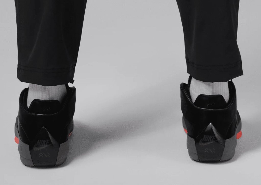 Back view of a person wearing black Nike sneakers with a gray and red sole, black pants, and white socks, against a plain background.