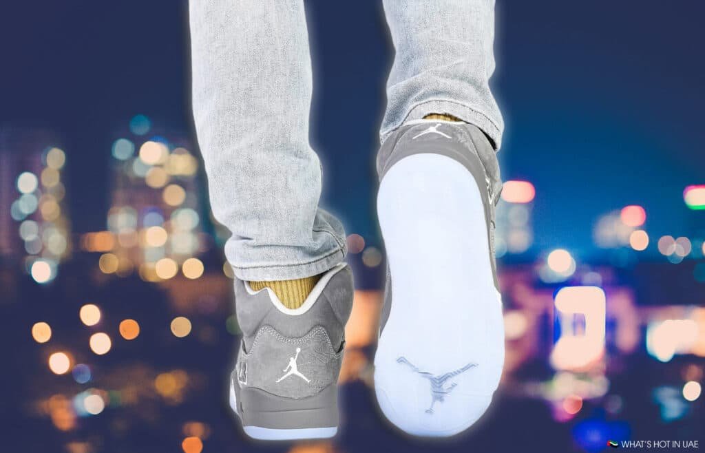 A person wearing light blue jeans and Air Jordan 5 Wolf Grey sneakers, featuring a white sole and logo, stands against a blurred cityscape with colorful bokeh lights.