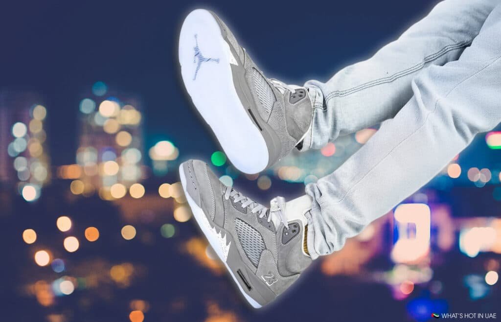 A pair of Air Jordan 5 Wolf Grey sneakers with sleek design details, worn with light blue jeans against a blurred city nightscape with colorful bokeh lights.